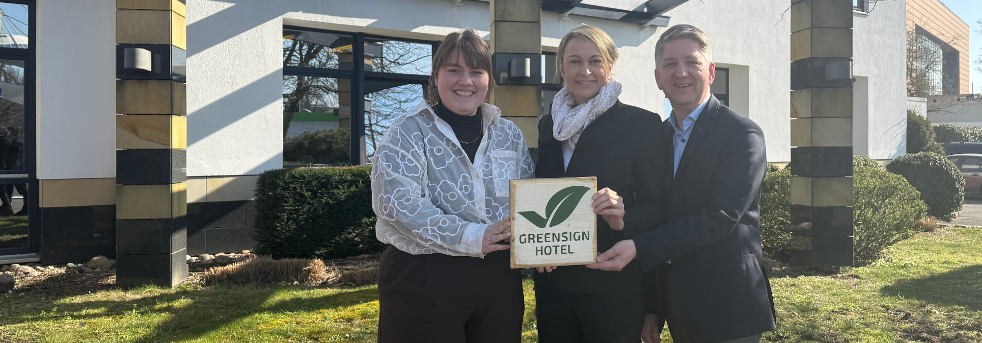 Drei Personen vor einem Gebäude. Die beiden Frauen gehören zur Familie Bock, der Hoteldirektor steht rechts. Sie halten ein Siegel für Nachhaltigkeit.