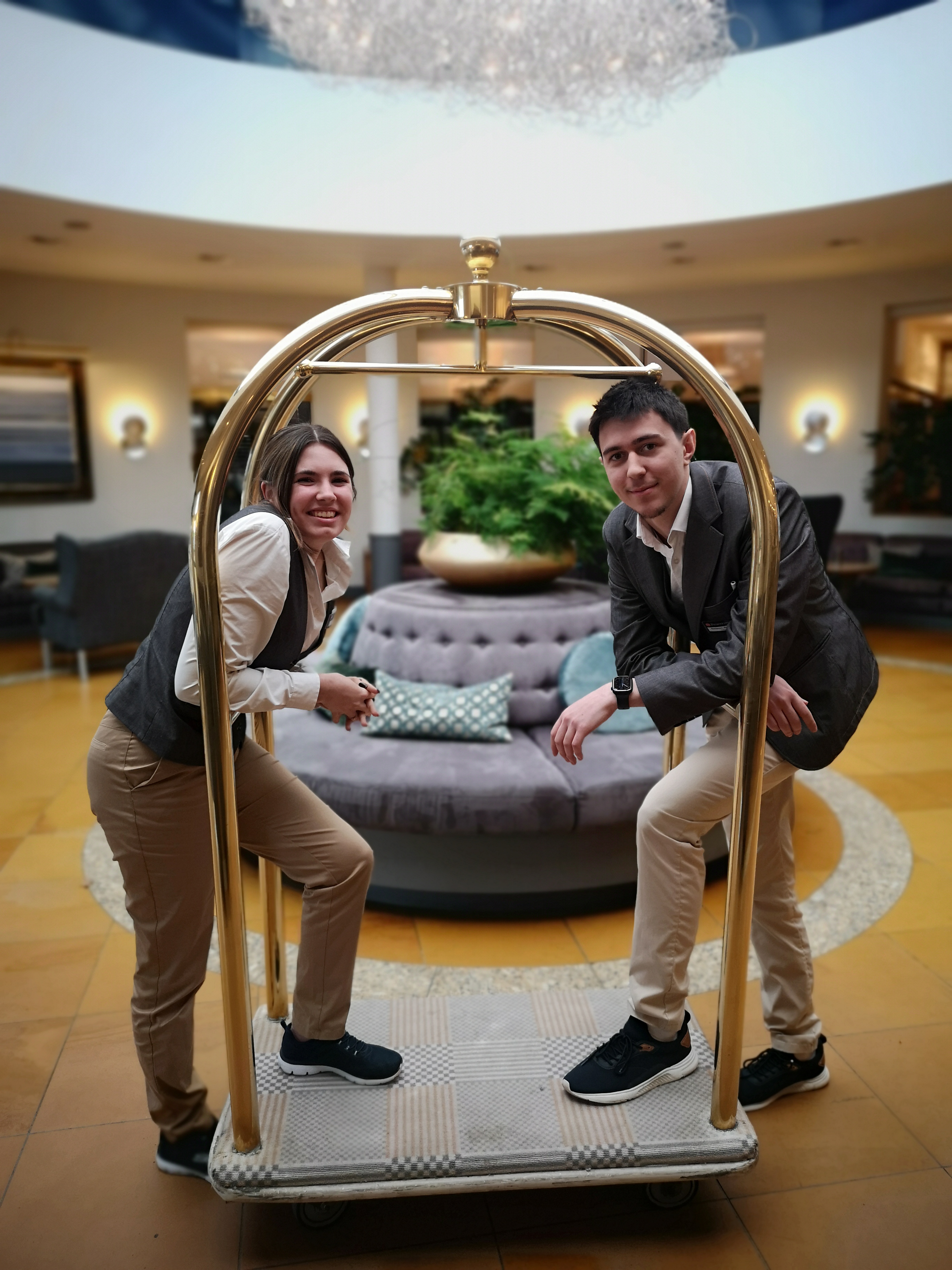 zwei Auszubildende auf Gep&auml;ckwagen, der in der Hotelhalle steht.